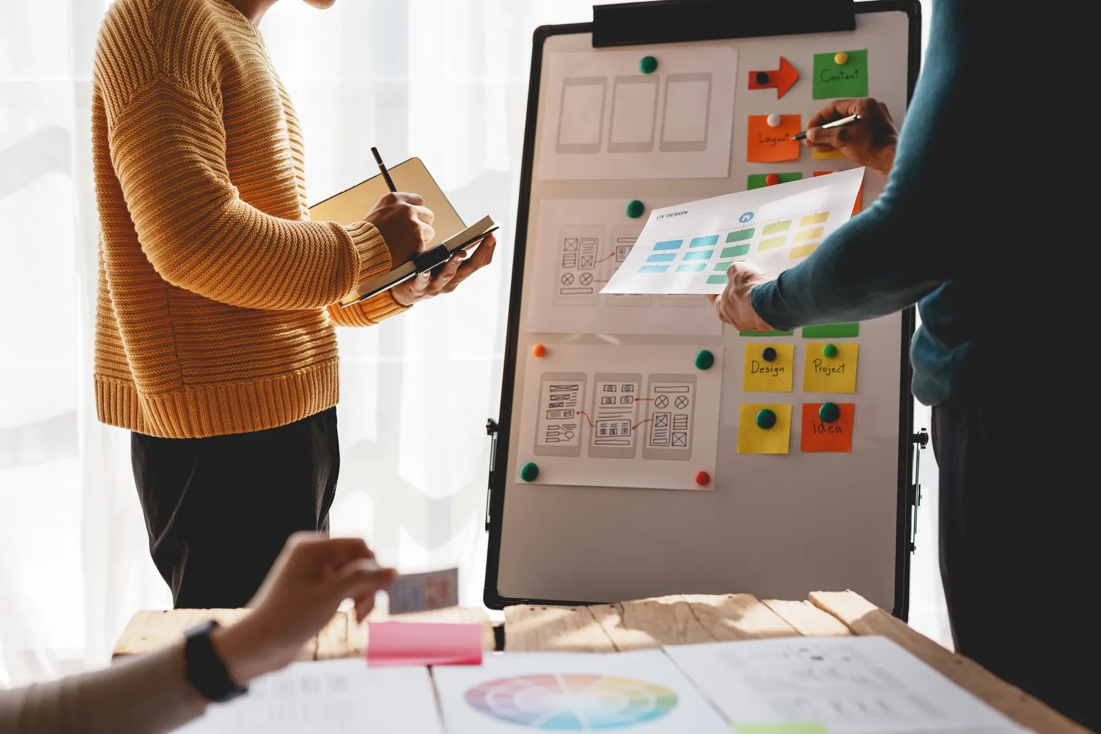 Two designers arranging wireframe cards on a whiteboard beside a printed colour wheel