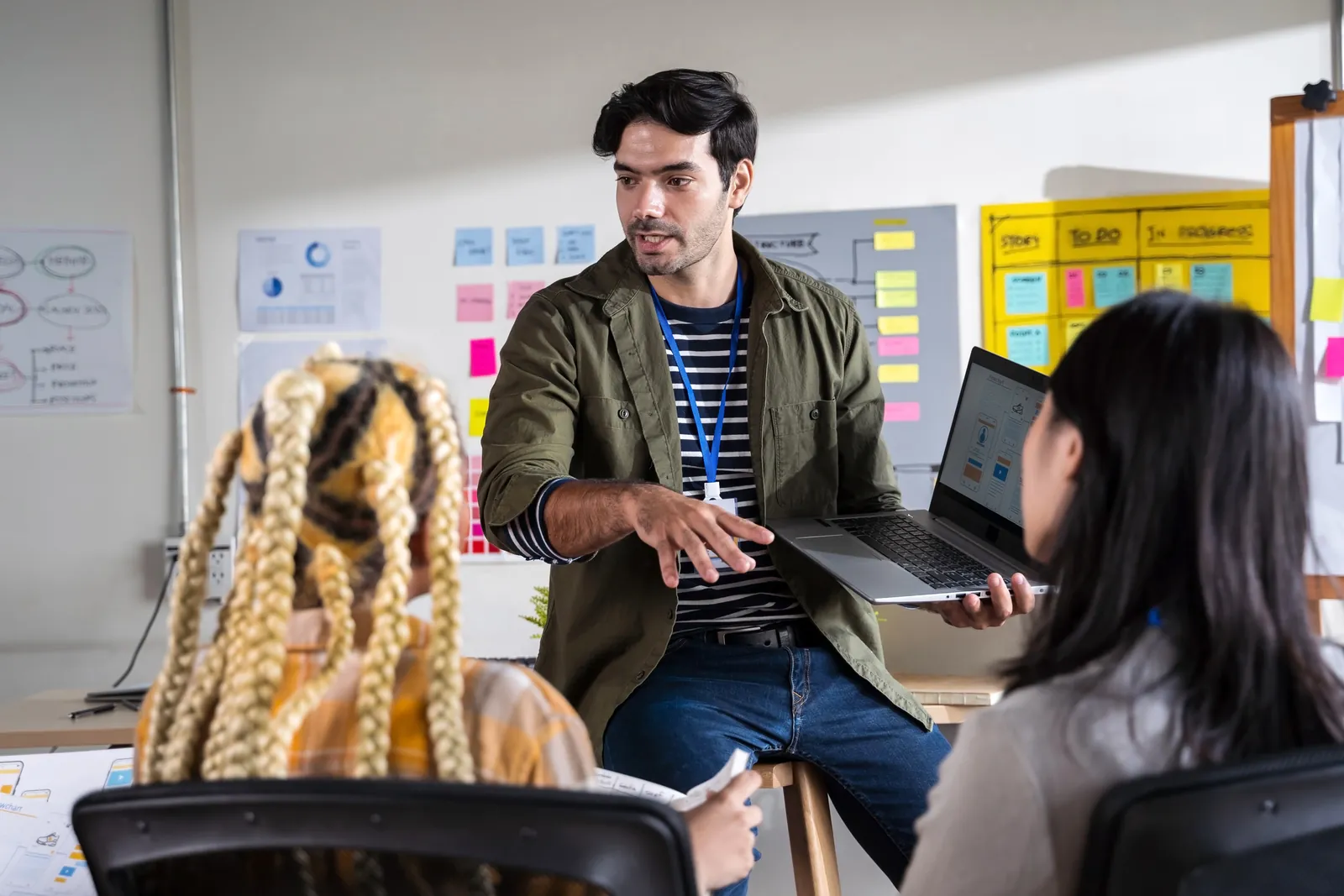 Designer holding a laptop presenting wireframes pinned on the wall to seated colleagues