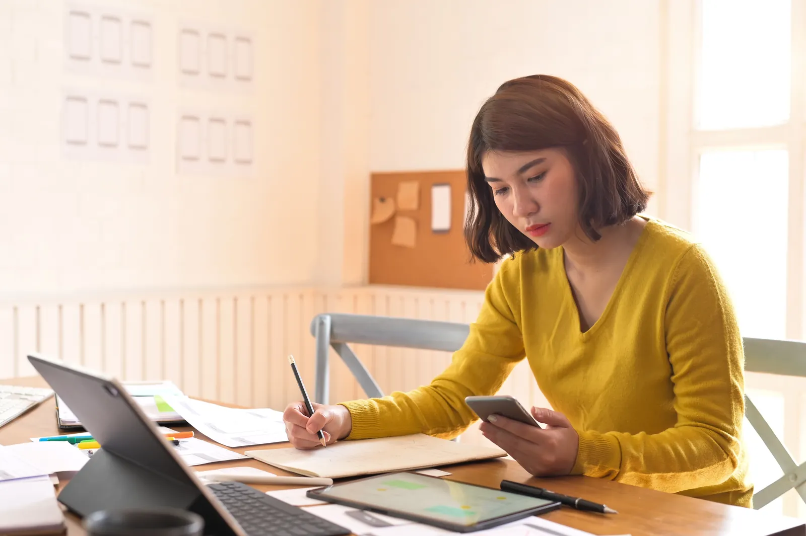 Designer in a yellow top taking notes from a phone beside a laptop
